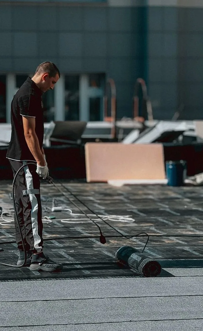 A construction worker applying a waterproofing coat on a rooftop with a roller during daytime.